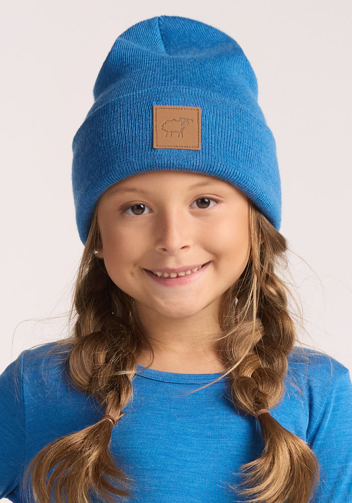 A young girl with braided hair smiles at the camera, wearing the Woolx Kids Baylor Beanie in Blue Razz and a matching blue shirt. The beanie features a brown patch. The background is plain and light-colored.