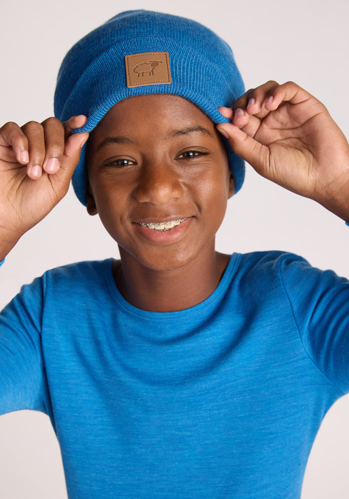 A smiling child wears the Woolx Kids Baylor Beanie in Blue Razz with a matching long-sleeve shirt, holding the beanie’s sides and looking directly at the camera.