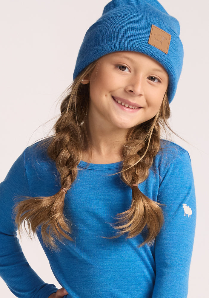 A young girl with long brown braids smiles while wearing the Woolx Kids Baylor Beanie - Blue Razz and a matching long-sleeve shirt with a small white sheep logo, posing against a light background.