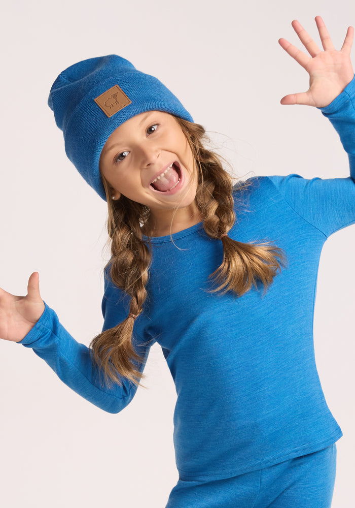 A young girl with long brown braided hair smiles widely, wearing the Woolx Kids Baylor Beanie in Blue Razz and a matching blue long-sleeve top, her hands raised playfully.