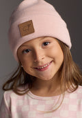 A young girl with long brown hair smiles while wearing the Woolx Kids Baylor Beanie - Little Ballerina, a light pink itch-free merino wool hat with a leather sheep logo patch. She pairs it with a pink and white checkered shirt against a gray background.