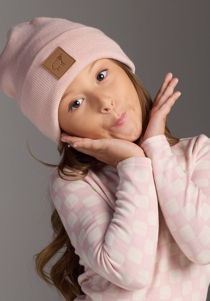 A young girl with long brown hair wears the Woolx Kids Baylor Beanie - Little Ballerina, a light pink merino wool winter hat, and a pink patterned shirt, playfully posing with her hands framing her face against a plain gray background.