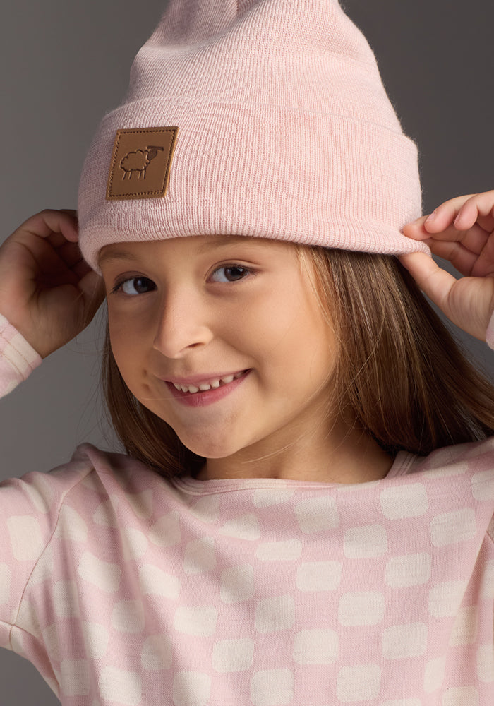 A young girl smiles as she adjusts her light pink Woolx Kids Baylor Beanie - Little Ballerina, made from itch-free merino wool with a sheep patch, pairing it with her square-patterned top against a plain background.