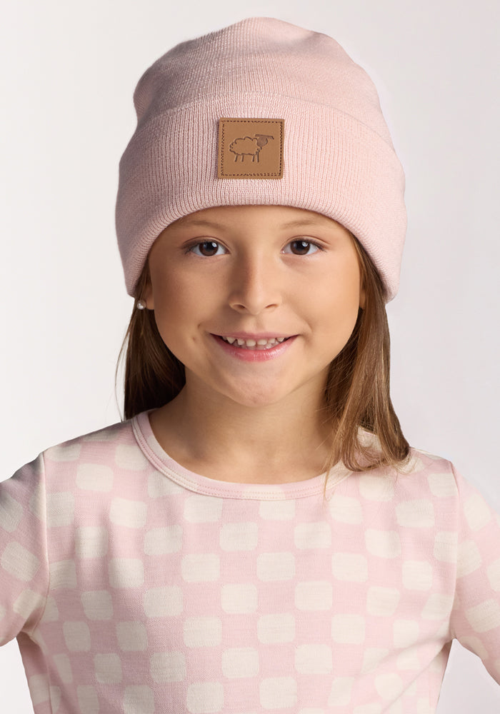 A smiling young girl wears the Woolx Kids Baylor Beanie - Little Ballerina, a pale pink, itch-free merino wool winter hat, with a light pink and white checkered shirt against a plain background—perfect cozy comfort for kids.
