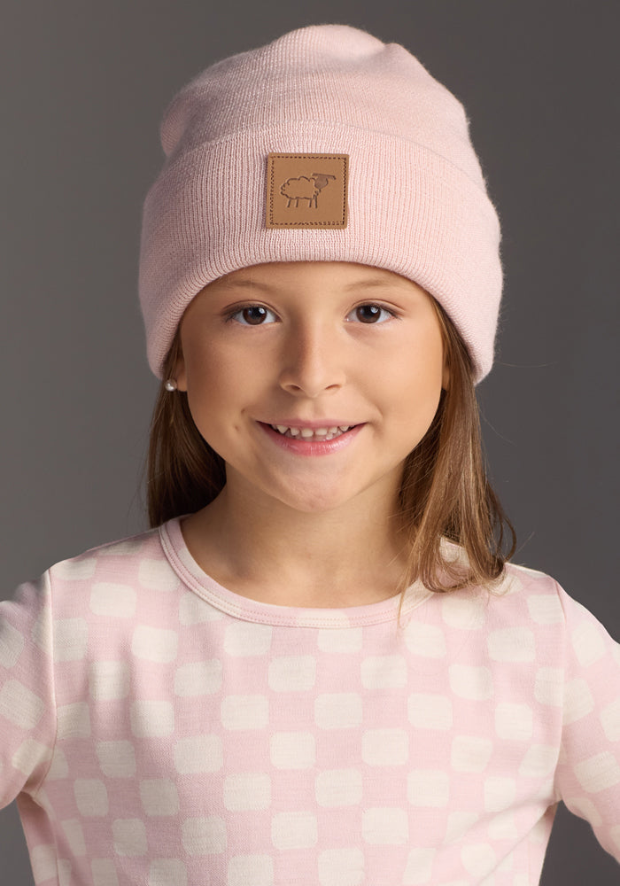 Smiling young girl wearing the Woolx Kids Baylor Beanie - Little Ballerina in light pink and a matching checkered shirt, standing in front of a plain gray background.