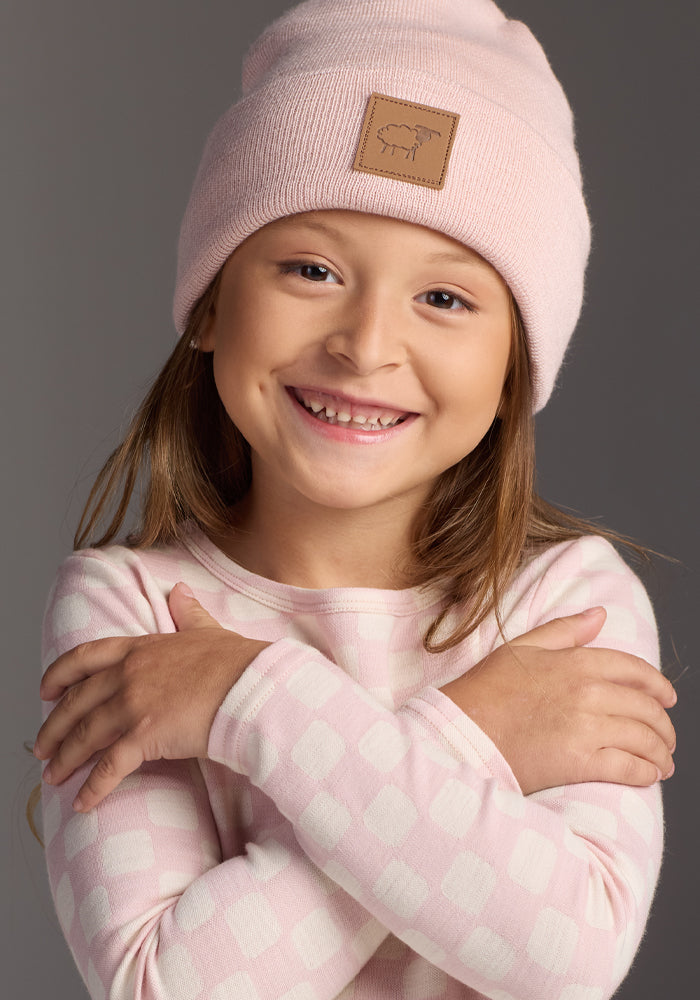 A young girl with long brown hair smiles, wearing the Woolx Kids Baylor Beanie in Little Ballerina (itch-free merino wool) and a light pink checkered long-sleeve shirt, standing with her arms crossed against a neutral background.