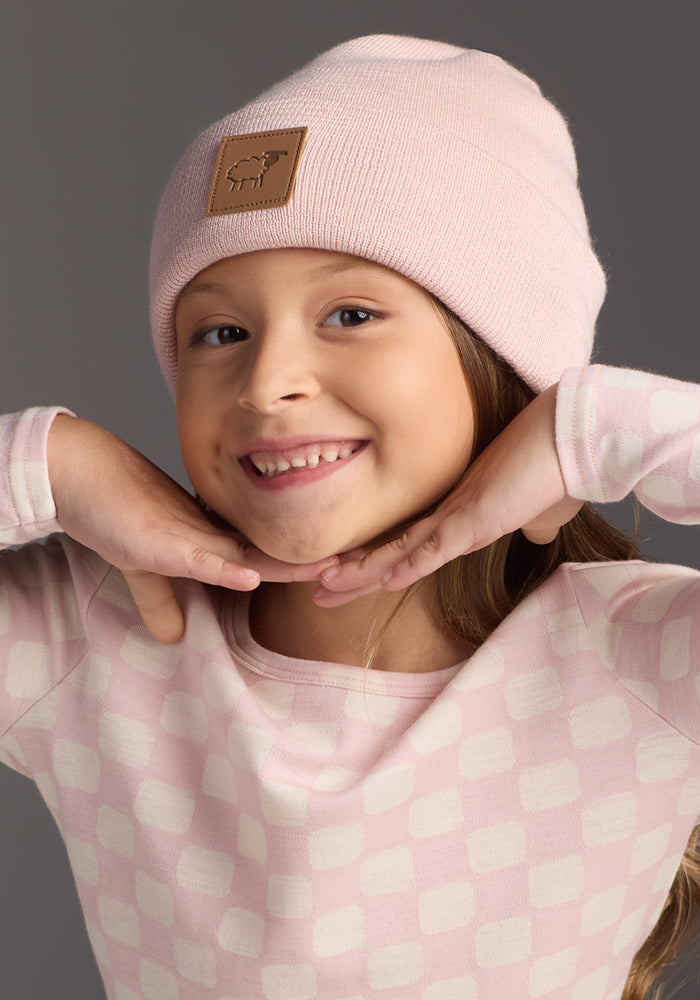 A young child smiles with their chin on their hands, wearing the Woolx Kids Baylor Beanie - Little Ballerina, a light pink merino wool winter hat that's soft and cozy against a neutral gray background.