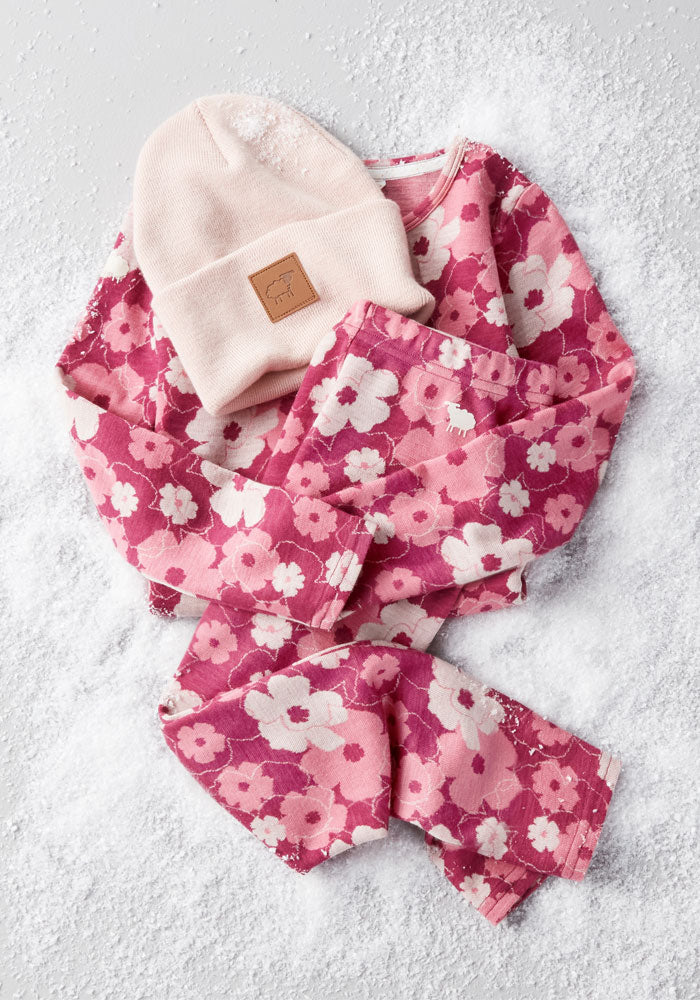 A pink floral long-sleeve shirt and matching pants are laid out on artificial snow, topped with Woolx’s Kids Baylor Beanie - Little Ballerina, an itch-free merino wool hat in light pink with a brown patch.