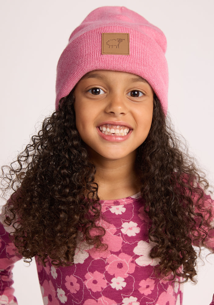 A young girl with long curly hair smiles at the camera, wearing the Woolx Kids Baylor Beanie - Pinky Promise and a pink floral-patterned shirt against a plain, light background.