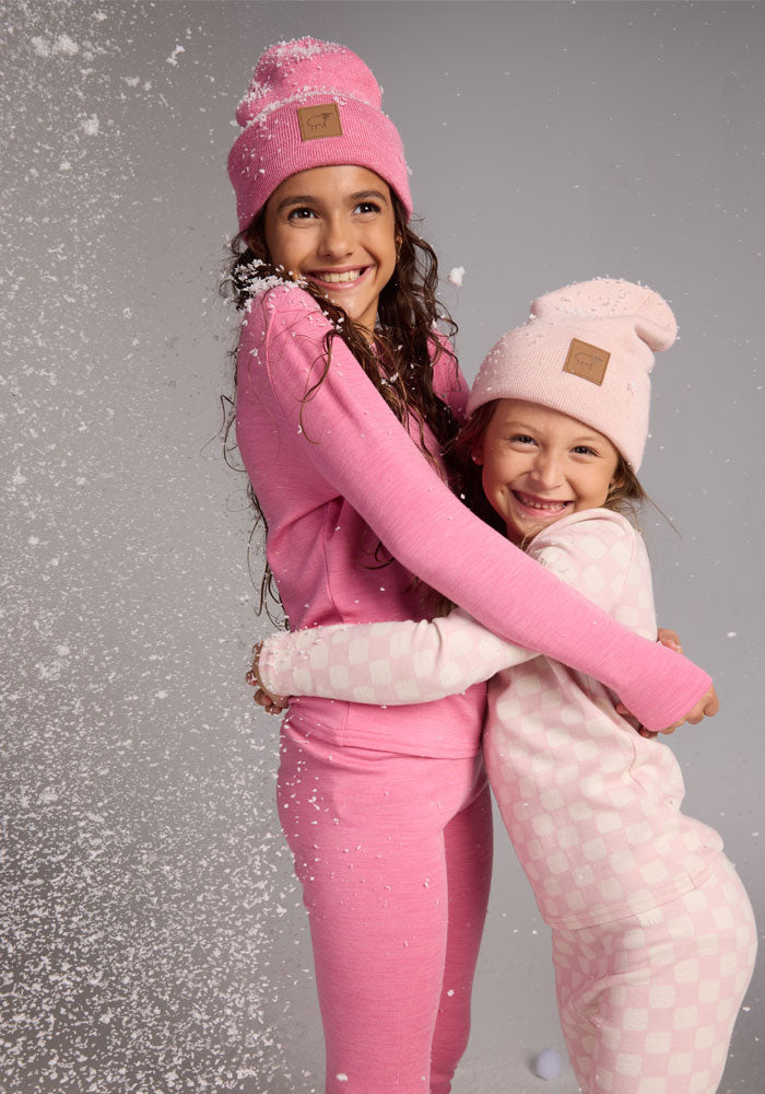 Two young girls in pink pajamas and matching Woolx Kids Baylor Beanie - Pinky Promise hats hug and smile as artificial snow falls around them, creating a playful winter scene.
