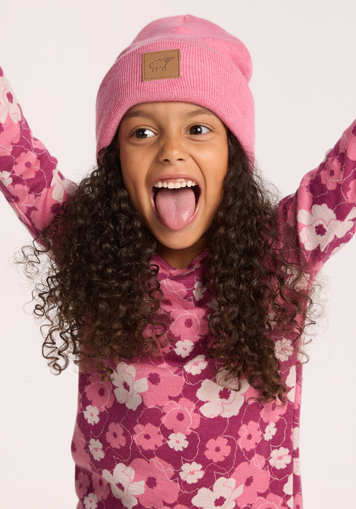A young girl with curly hair, wearing a pink floral shirt and the Woolx Kids Baylor Beanie - Pinky Promise, smiles widely with her mouth open and arms raised, sticking out her tongue against a plain light background.
