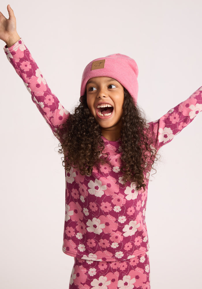 A young girl with curly hair, wearing the Woolx Kids Baylor Beanie - Pinky Promise and a pink floral long-sleeve shirt, smiles joyfully with her arms raised against a plain light background.