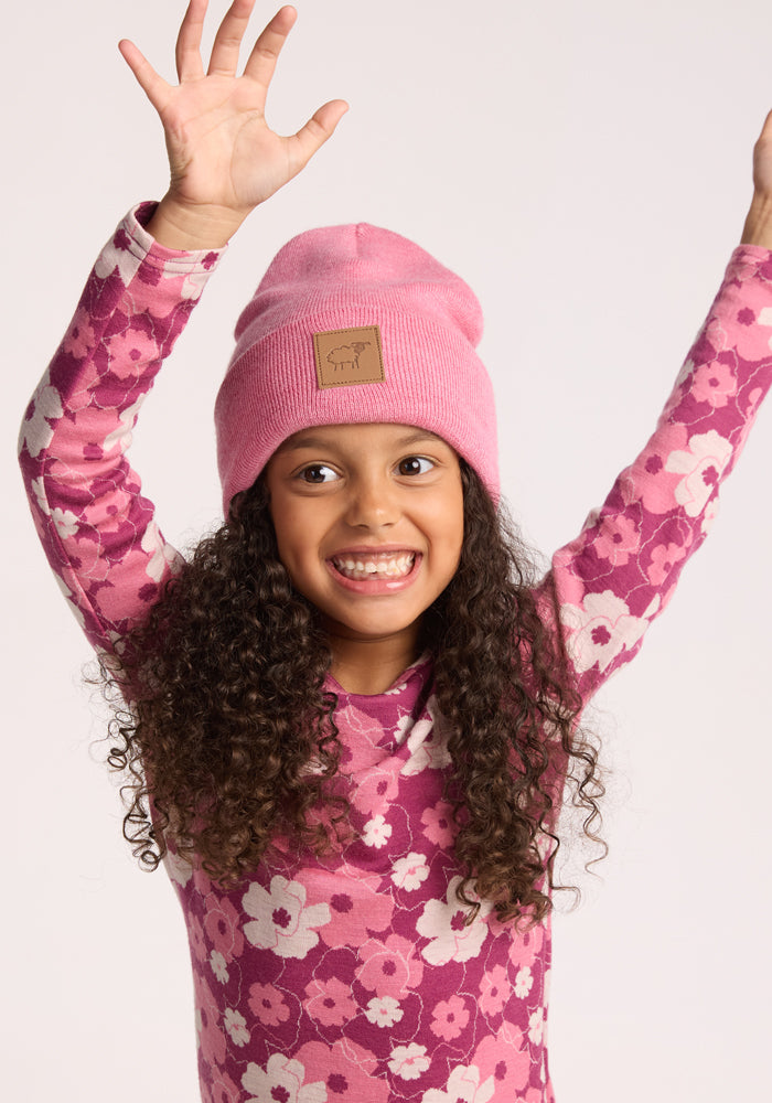 A smiling young girl with long curly hair raises her arms cheerfully, wearing the Woolx Kids Baylor Beanie - Pinky Promise and a pink floral long-sleeve shirt against a light background.