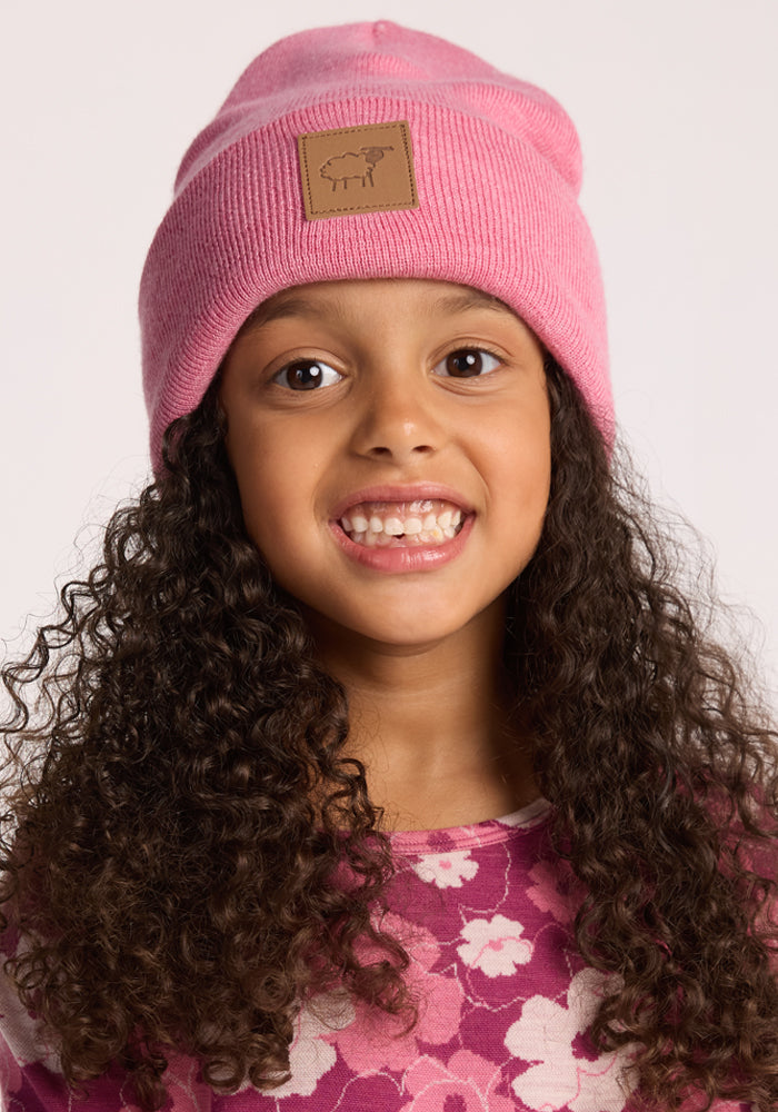 A smiling young girl with long, curly brown hair wears the Woolx Kids Baylor Beanie in Pinky Promise and a pink floral-patterned shirt, standing against a plain light background.