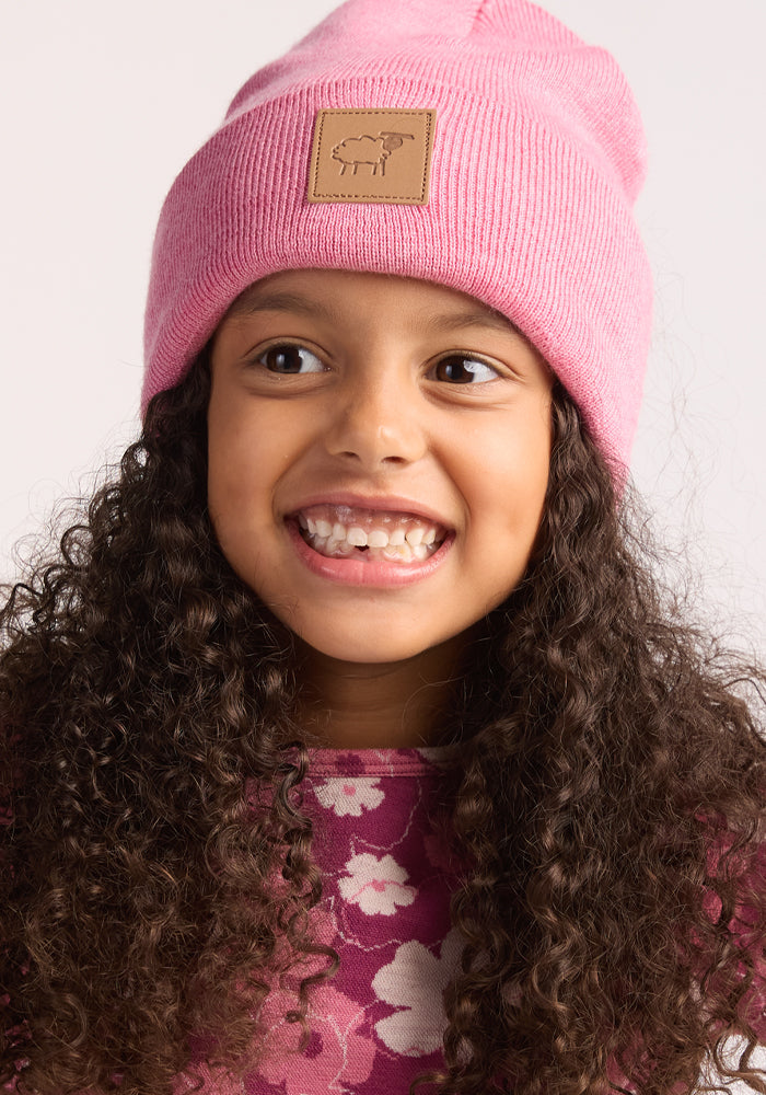 A young girl with curly hair smiles, showing missing front teeth. She wears a Woolx Kids Baylor Beanie - Pinky Promise and a pink floral-patterned shirt.
