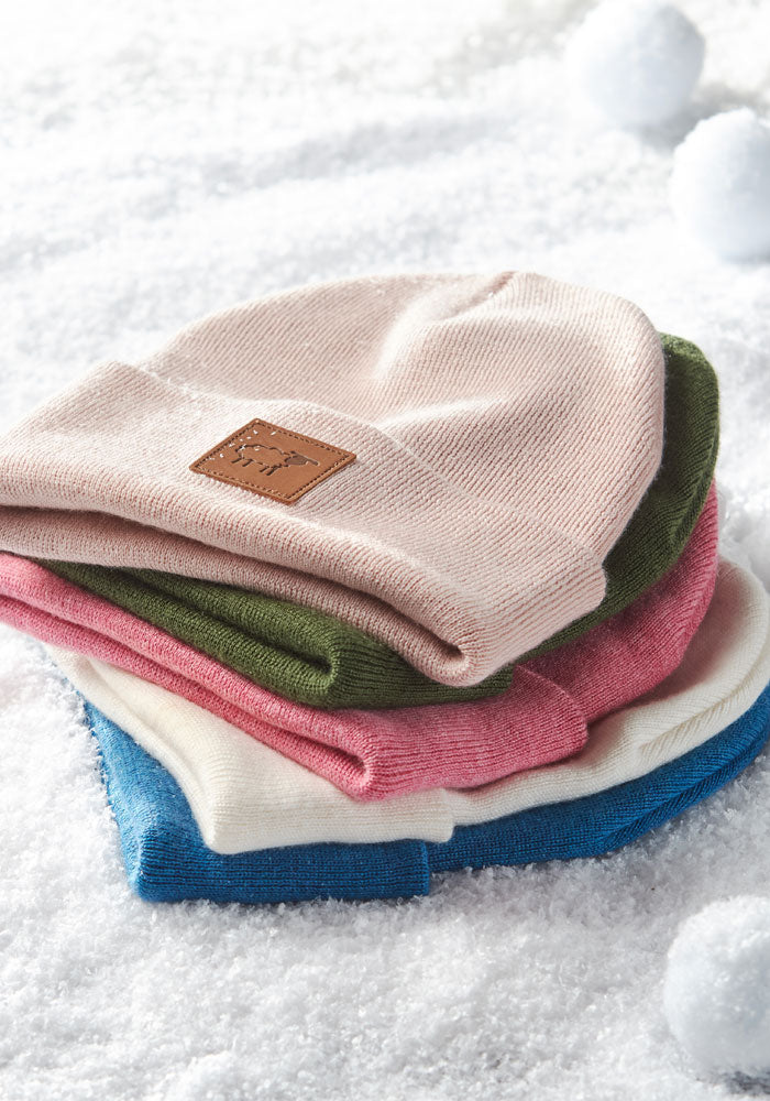 Five stretchy Woolx Kids Baylor Beanie - Pinky Promise winter hats in pastel colors—pink, green, white, red, and blue—are stacked on snow with snowballs behind. The top hat has a brown leather patch.