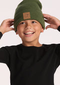 A smiling child wears a Woolx Kids Baylor Beanie - Turtle Shell, featuring ear coverage and a brown patch, with a black long-sleeve shirt, holding the beanie's sides against a plain light background.