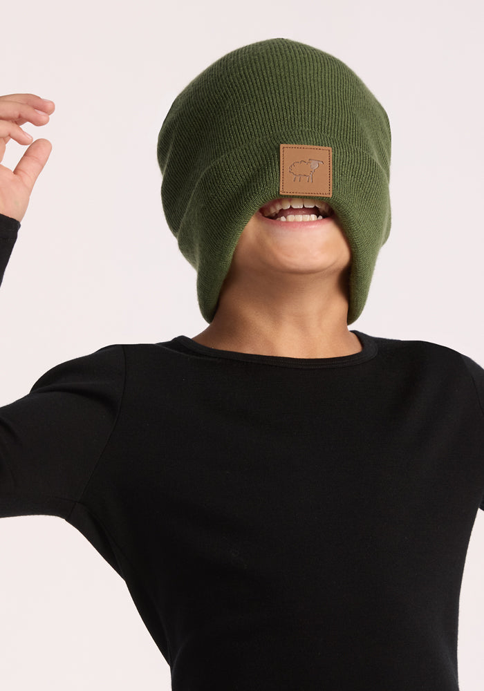 A child wearing a black shirt and the Woolx Kids Baylor Beanie - Turtle Shell pulled over their face smiles playfully, raising their arms for full ear coverage against a plain background.