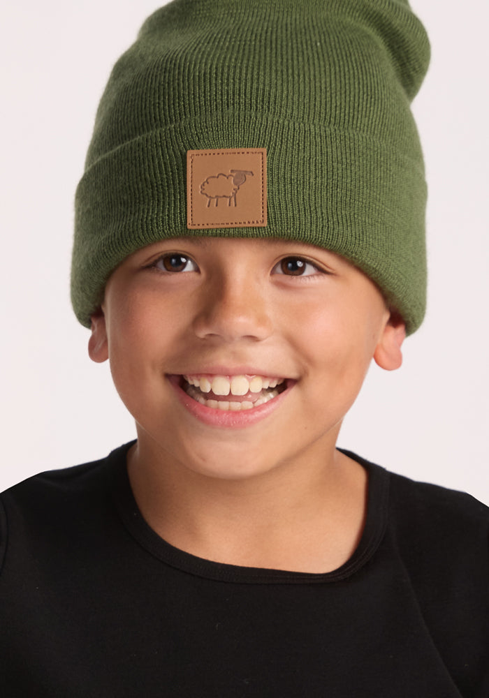 A smiling child wears the Woolx Kids Baylor Beanie in Turtle Shell, a green merino wool hat with ear coverage and a brown patch featuring a simple sheep illustration, paired with a black shirt against a plain light background.