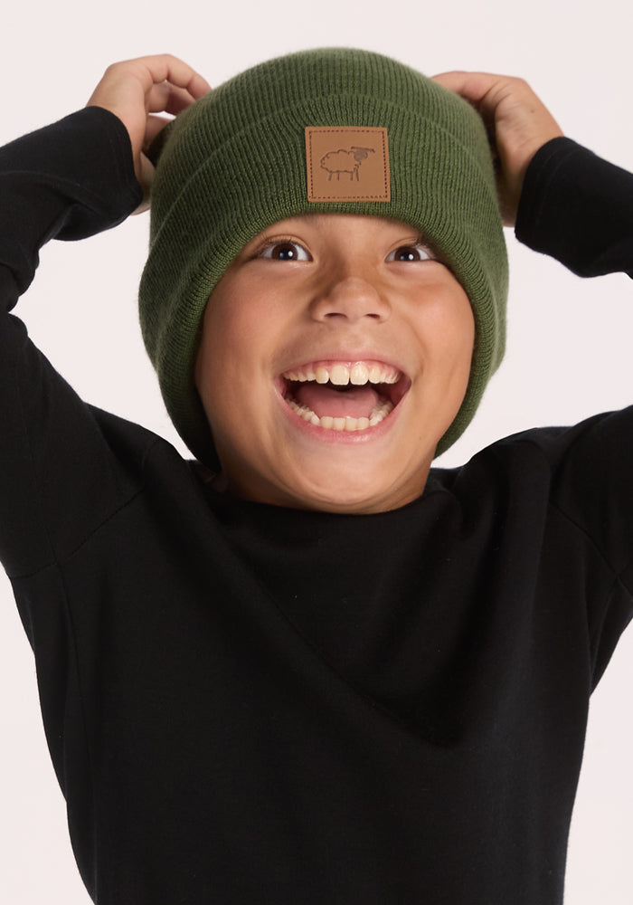 A smiling child in a Woolx Kids Baylor Beanie - Turtle Shell, featuring a patch and stretch fit for cozy ear coverage, wears a black long-sleeve shirt and raises their hands to their head, looking excited against a plain background.
