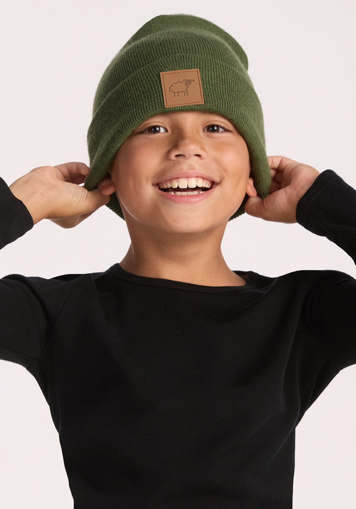 A smiling child wears the Woolx Kids Baylor Beanie - Turtle Shell, featuring a brown patch and stretch fit for cozy ear coverage, paired with a black long-sleeve shirt, against a plain light background.