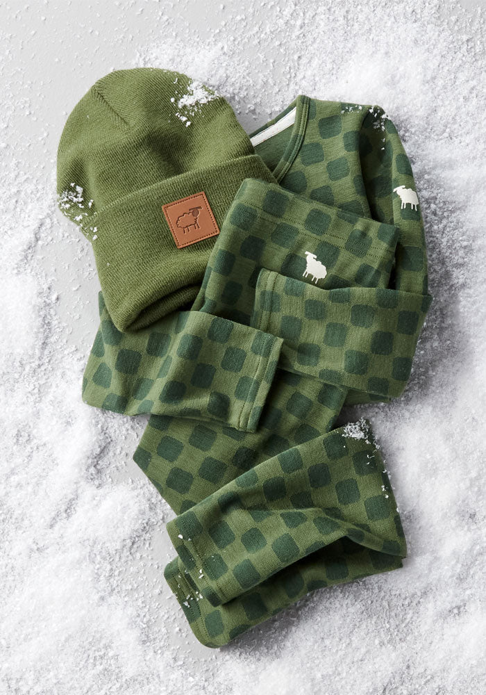 A green checkered pajama set with a sheep logo is paired with the Woolx Kids Baylor Beanie - Turtle Shell, a green merino wool hat with full ear coverage and brown patch, both laid on snow-dusted ground.