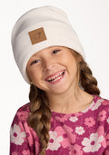A young girl with long braided hair smiles, wearing the Woolx Kids Baylor Beanie in Whipped Cream with a brown patch and a pink floral shirt, against a plain white background.
