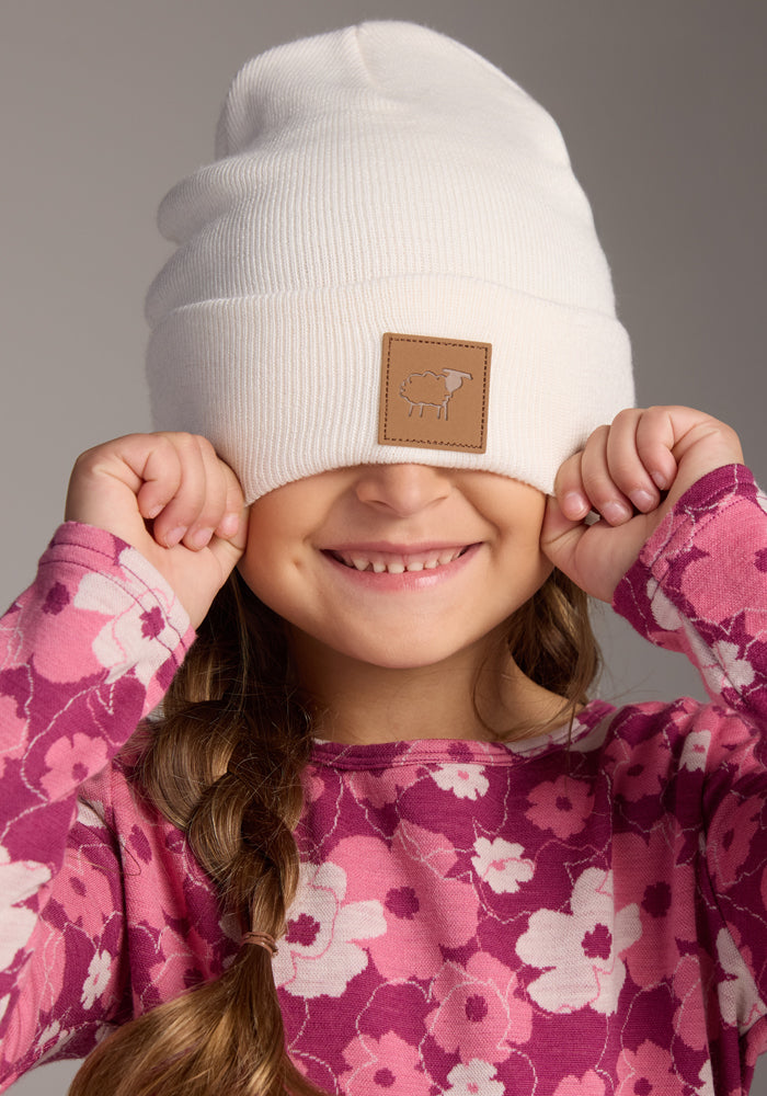 A smiling child in a pink floral shirt pulls the Woolx Kids Baylor Beanie in Whipped Cream, featuring a tan patch, down over their eyes. The cozy wool winter hat pops against the plain, neutral background.