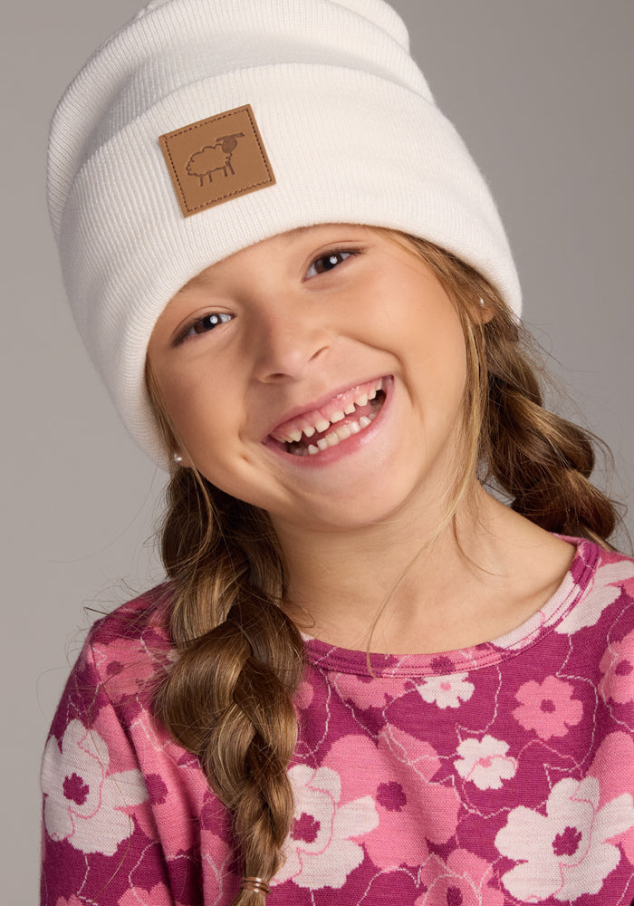 A young girl with braided hair smiles, wearing the Woolx Kids Baylor Beanie in Whipped Cream and a pink floral shirt, set against a neutral background.