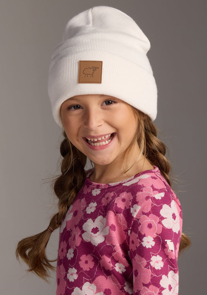 A smiling young girl with two braids wears the Woolx Kids Baylor Beanie in Whipped Cream and a pink floral shirt, standing against a plain gray background.