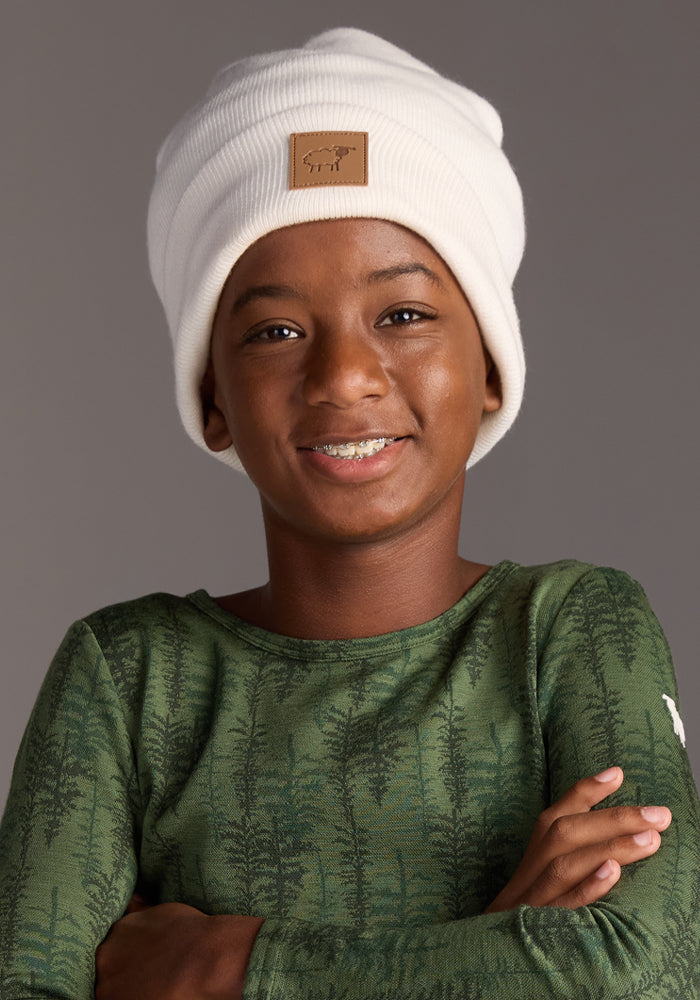 A smiling child in the Woolx Kids Baylor Beanie - Whipped Cream and a green tree-print long-sleeve shirt poses arms crossed against a plain gray background.
