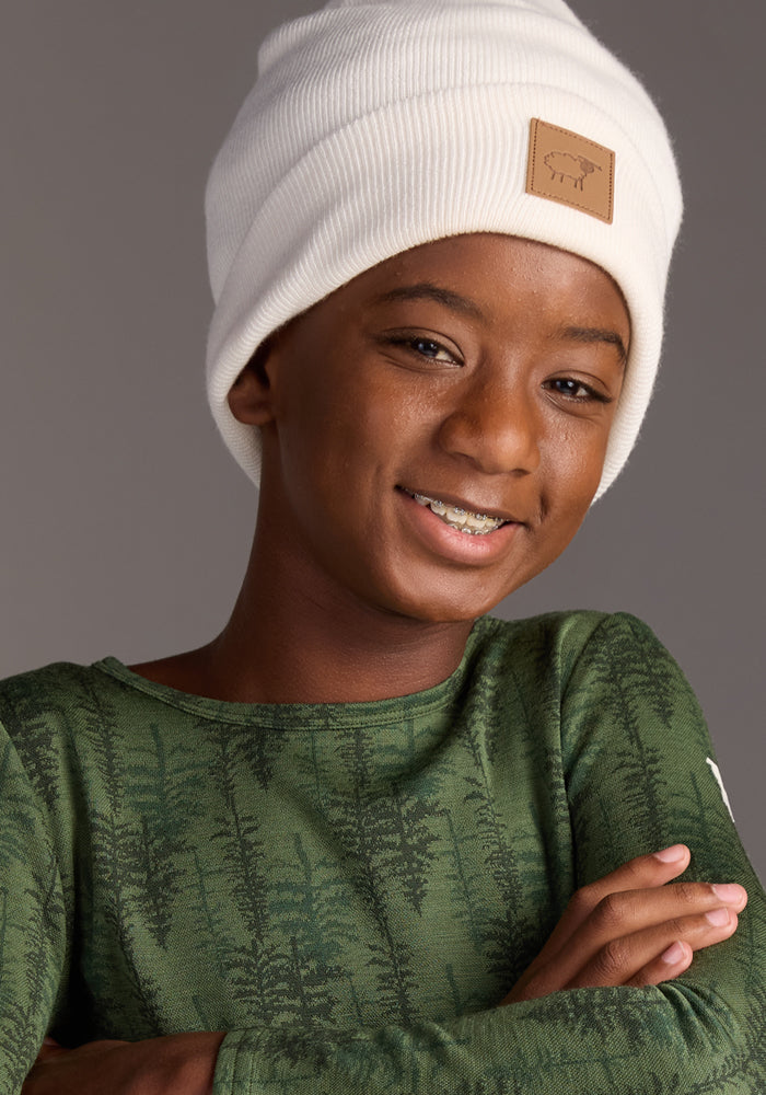 A smiling child with dark skin wears the Woolx Kids Baylor Beanie in Whipped Cream and a green long-sleeve shirt with a tree pattern, standing with arms crossed against a plain gray background.