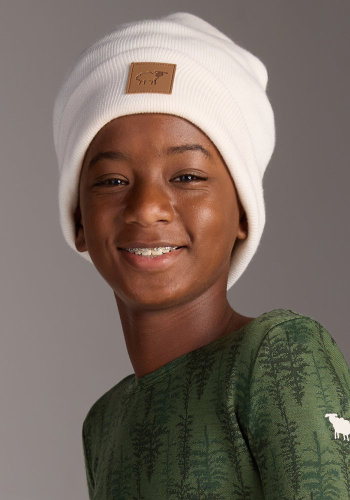 A smiling child wears the Woolx Kids Baylor Beanie in Whipped Cream and a green tree-patterned shirt, standing against a neutral background.