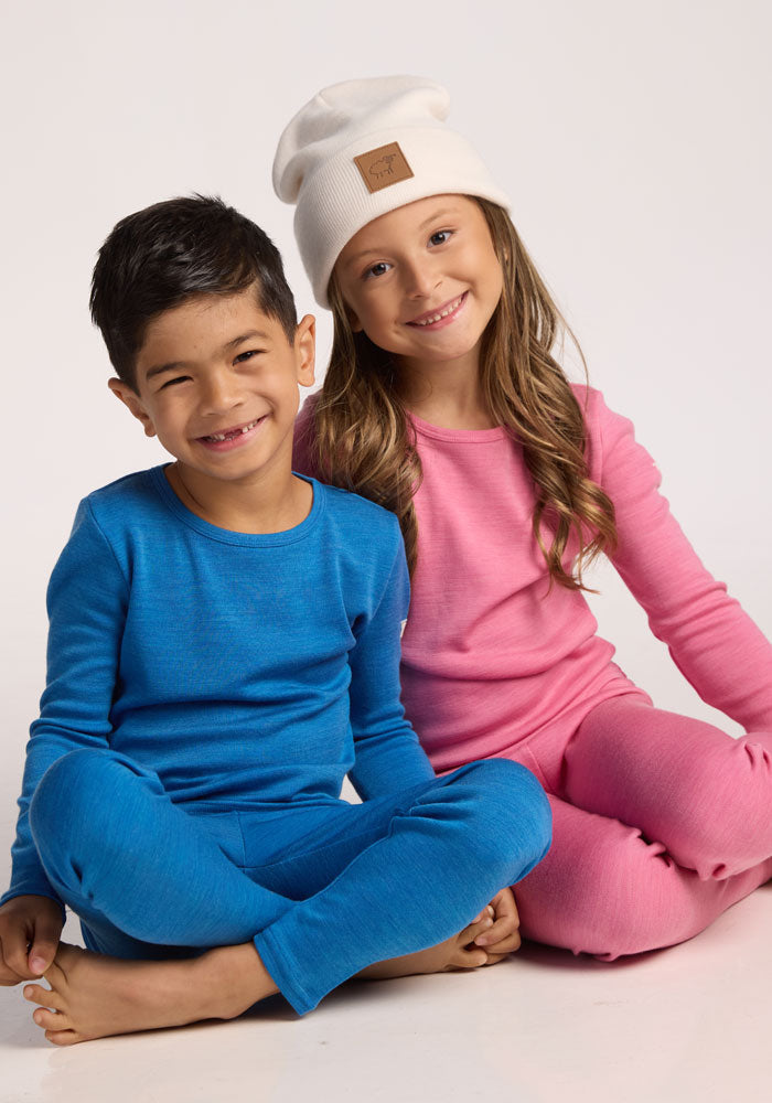 A young boy in blue pajamas, a young girl in pink pajamas, and a cozy Woolx Kids Baylor Beanie in Whipped Cream sit on the floor smiling against a plain white background.