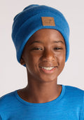 A smiling boy wears the Woolx Kids Baylor Beanie in Blue Razz with a tan patch, paired with a blue shirt, standing against a plain light background.