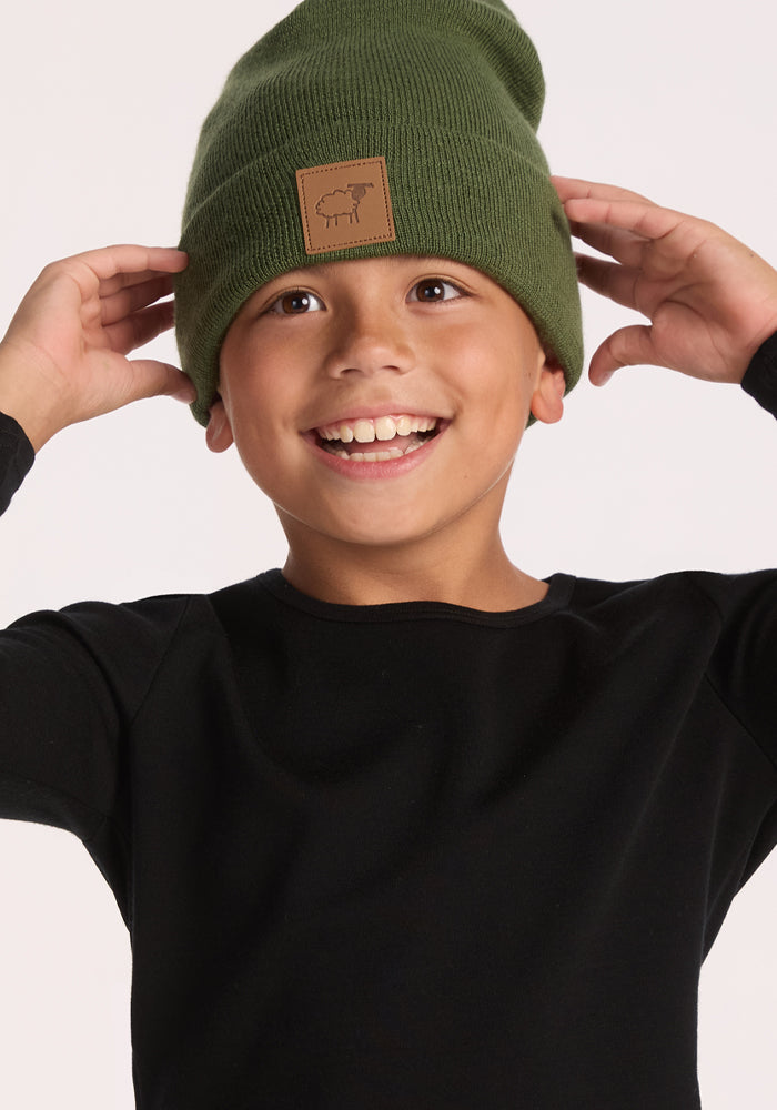 A smiling child wears a Woolx Kids Baylor Beanie - Turtle Shell, featuring ear coverage and a brown patch, with a black long-sleeve shirt, holding the beanie's sides against a plain light background.