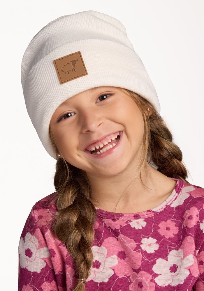 A young girl with long braided hair smiles, wearing the Woolx Kids Baylor Beanie in Whipped Cream with a brown patch and a pink floral shirt, against a plain white background.