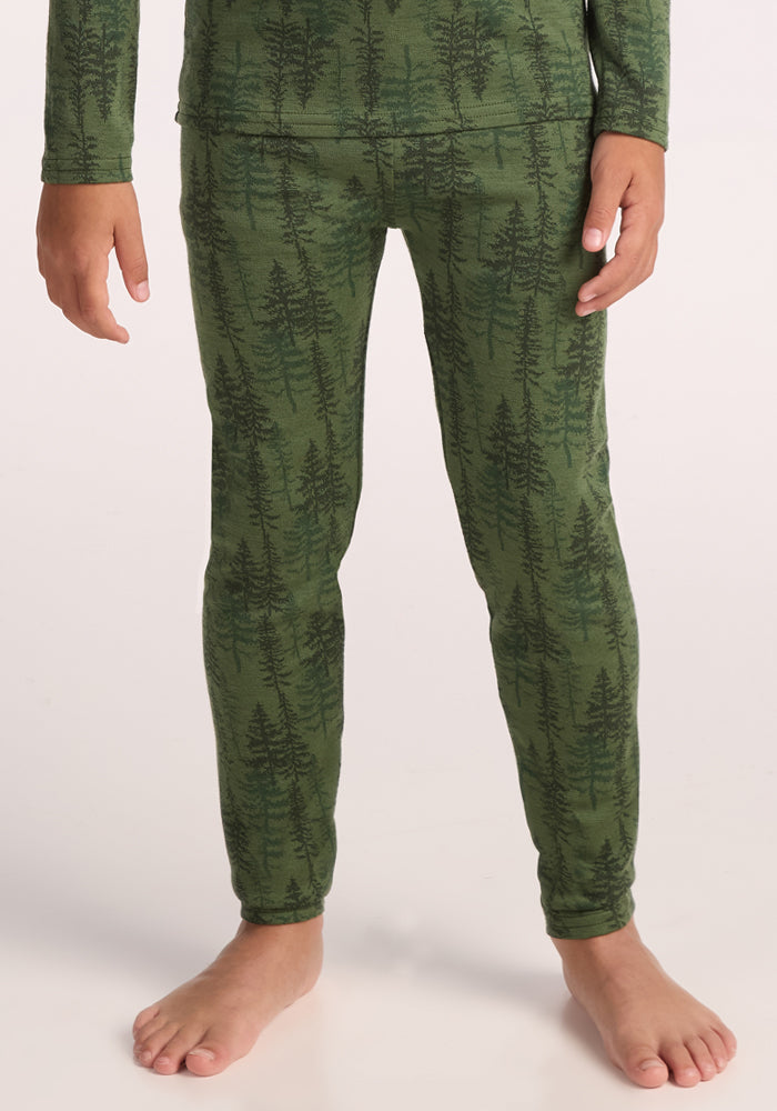 A child stands barefoot in Woolx Kids Explorer Base Layer Bottoms - Forest Friends, cozy green pajama pants with a dark tree pattern, ideal as a warm baselayer or comfy kids ski wear, set against a light background. | Rafael is wearing a size MED.