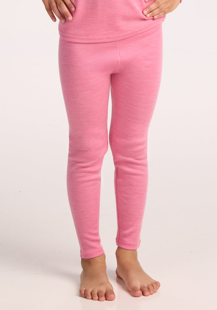 A child stands barefoot in Woolx Kids Explorer Base Layer Bottoms in Pinky Promise, paired with a matching merino wool long-sleeve top. Hands rest on hips against a plain, light background. | Penny is wearing a size SML.