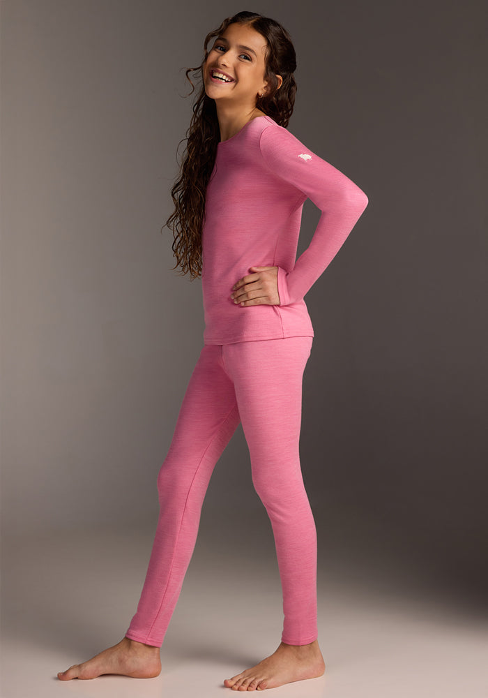 A smiling child stands barefoot on a gray background, wearing Woolx Kids Explorer Base Layer Bottoms - Pinky Promise—a cozy merino wool base layer, perfect for cooler days.
