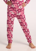 A child stands barefoot, wearing Woolx Kids Explorer Base Layer Bottoms in Rosie Posie under pink and white floral pajamas. The pajamas have long sleeves, and the background is plain and light-colored with the child’s hands on their hips. | Penny is wearing a size SML.