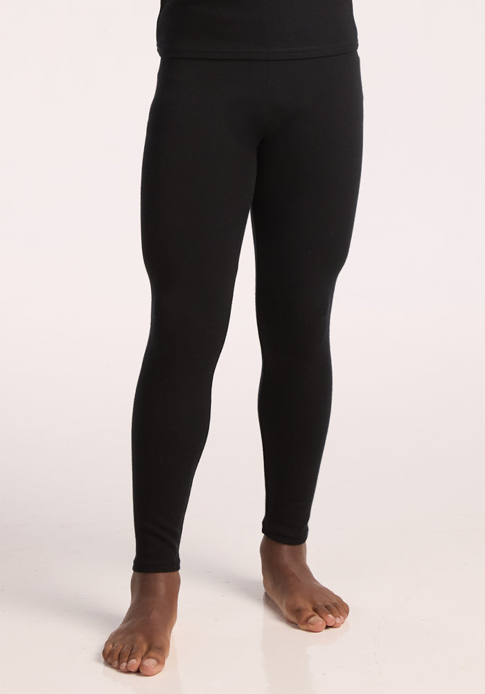 A child stands barefoot in Woolx Kids Explorer Base Layer Bottoms in black, against a light background. The image is cropped from the waist to the ankles. | Banyan is wearing a size LRG.