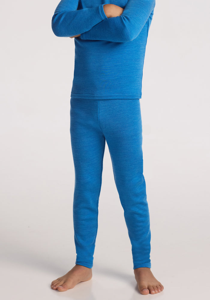 A child stands barefoot on a white background, arms crossed and head out of frame, wearing Woolx Kids Explorer Base Layer Bottoms in Blue Razz and a matching long-sleeved merino wool top. | Leo is wearing a size XSM.