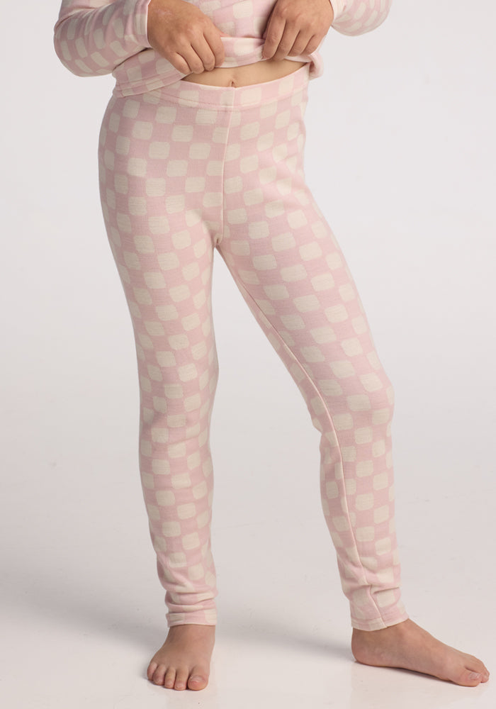 A child stands barefoot on a white background, wearing Woolx Kids Explorer Base Layer Bottoms in Hopscotch Pink checkered print and resting one hand on their waistband. | Aria is wearing a size SML.