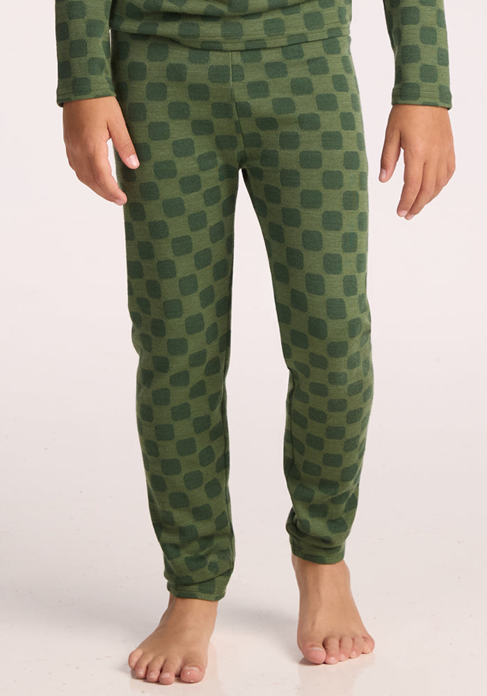 A child wearing Woolx Kids Explorer Base Layer Bottoms in Frog Hop, featuring a dark green square pattern, stands barefoot on a light surface. These cozy baselayer pants offer warmth and comfort. The upper body is partially visible. | Rafael is wearing a size MED.