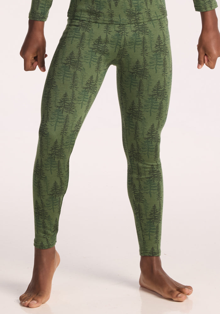 A child stands barefoot, shown from the shoulders down against a pale background, wearing Woolx Kids Explorer Base Layer Bottoms in the Forest Friends color. Their fists are gently clenched at their sides. | Banyan is wearing a size LRG.