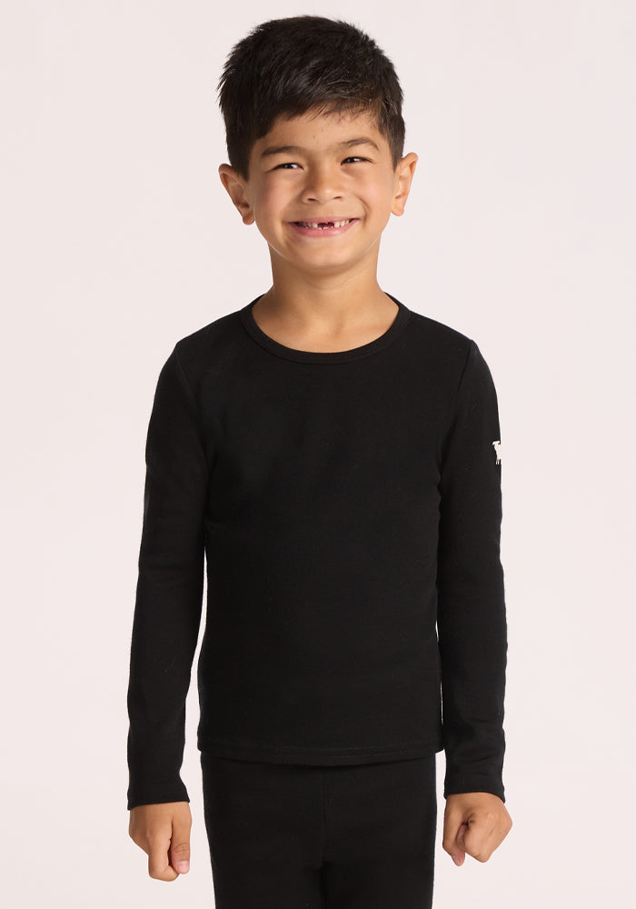 A young boy with dark hair smiles against a plain light background, wearing the Woolx Kids Explorer Base Layer Top in black, crafted from moisture-wicking Australian Merino wool, paired with matching black pants. | Leo is wearing a size XSM.