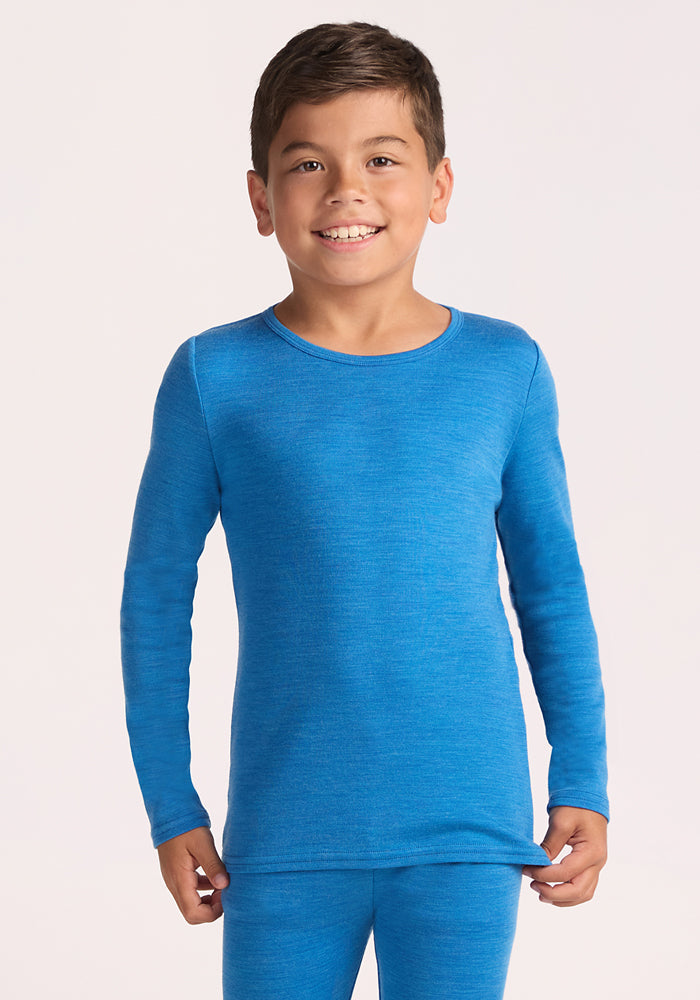 A young boy smiles at the camera wearing the Woolx Kids Explorer Base Layer Top in Blue Razz, made from soft Australian Merino wool. He stands against a plain light background, holding the edge of his shirt with one hand. | Rafael is wearing a size MED.
