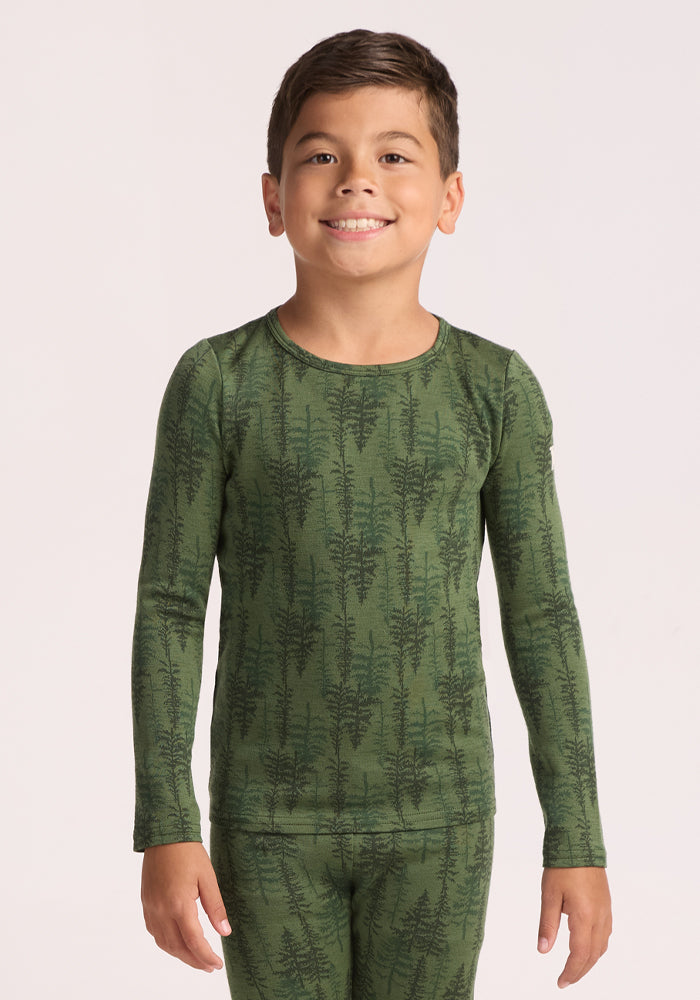 A young boy with short brown hair smiles at the camera, wearing the Woolx Kids Explorer Base Layer Top in Forest Friends, a moisture-wicking merino wool design with a dark tree pattern, standing against a plain light background. | Rafael is wearing a size MED.