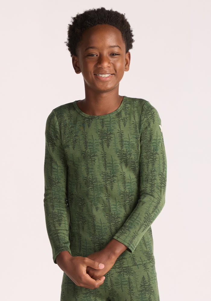 A smiling child with short curly hair wears the Woolx Kids Explorer Base Layer Top - Forest Friends, a green merino wool base layer with a subtle tree pattern, set against a plain light background. | Banyan is wearing a size LRG.